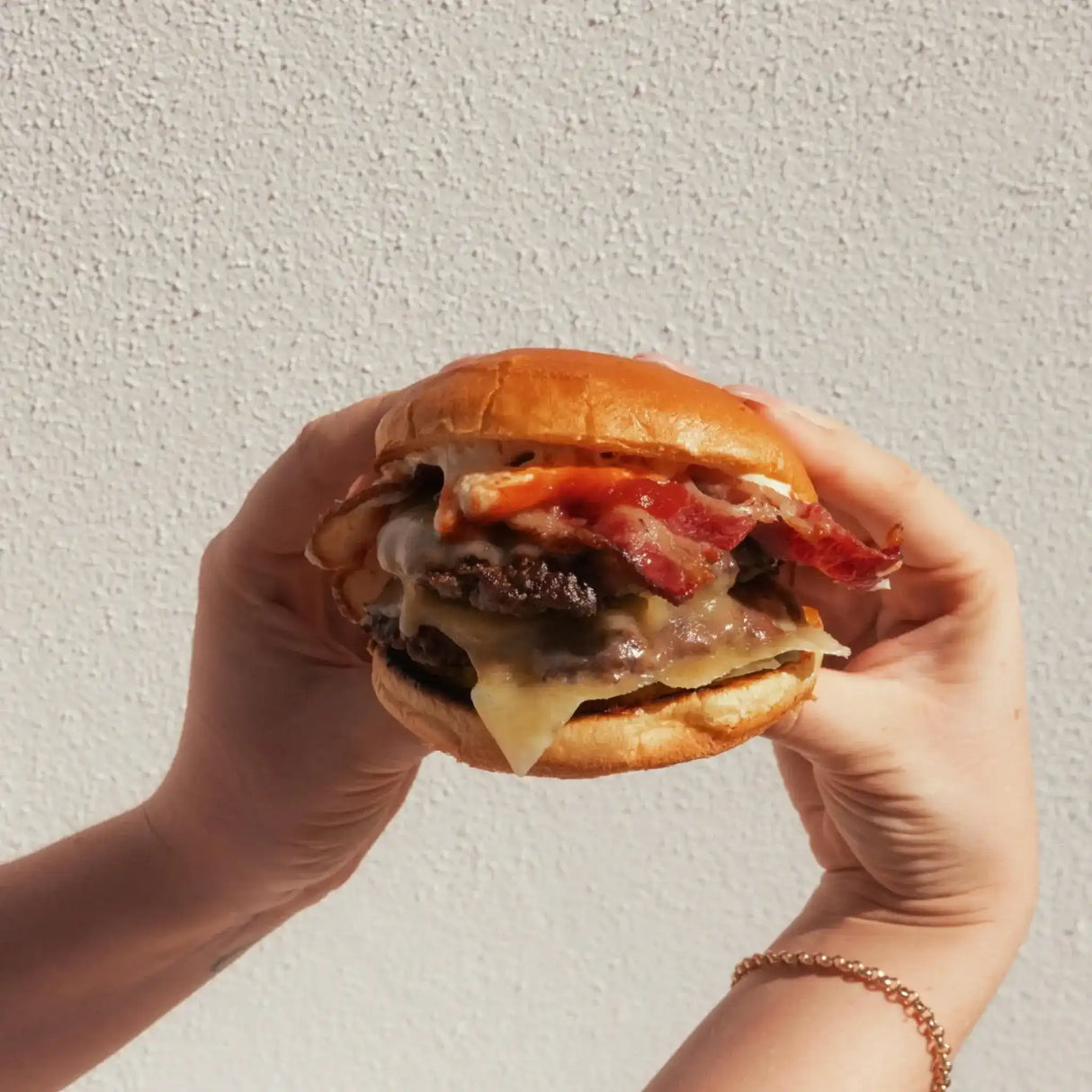 Juicy beef burger with melted cheese and crispy bacon, held in hands against textured wall