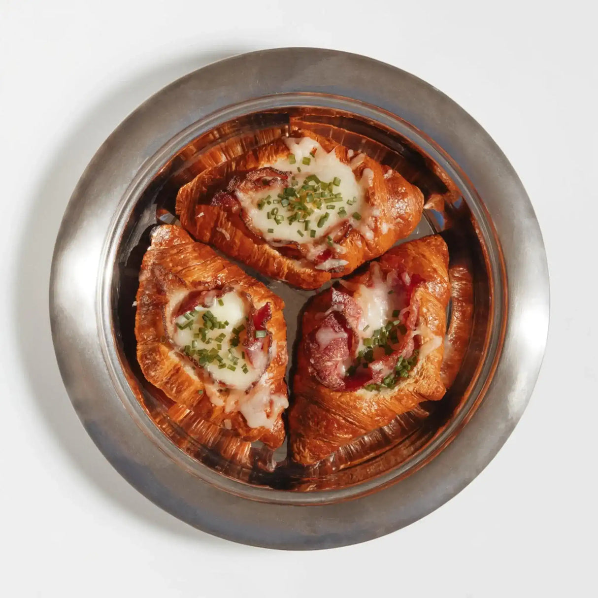 Three savory croissants with melted cheese, bacon, and chives on a metal plate