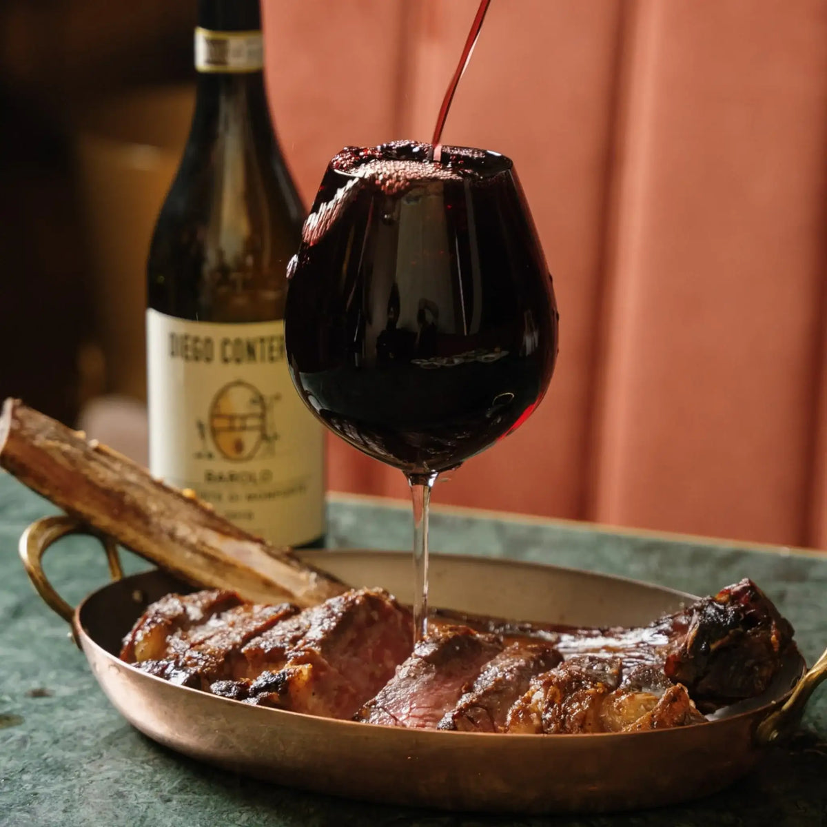 Sliced grilled steak in copper pan with red wine being poured, bottle in background
