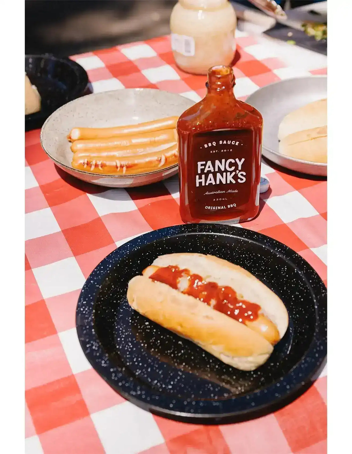Hot dog with BBQ sauce, sausage rolls, and Fancy Hank's BBQ sauce bottle on picnic table