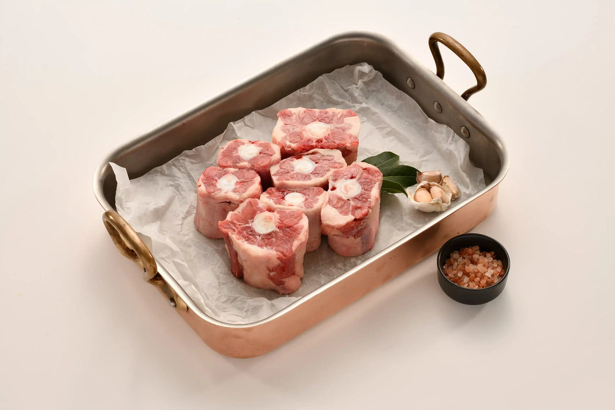 Raw oxtail pieces in a roasting pan with bay leaves, garlic, and pink salt on white background.