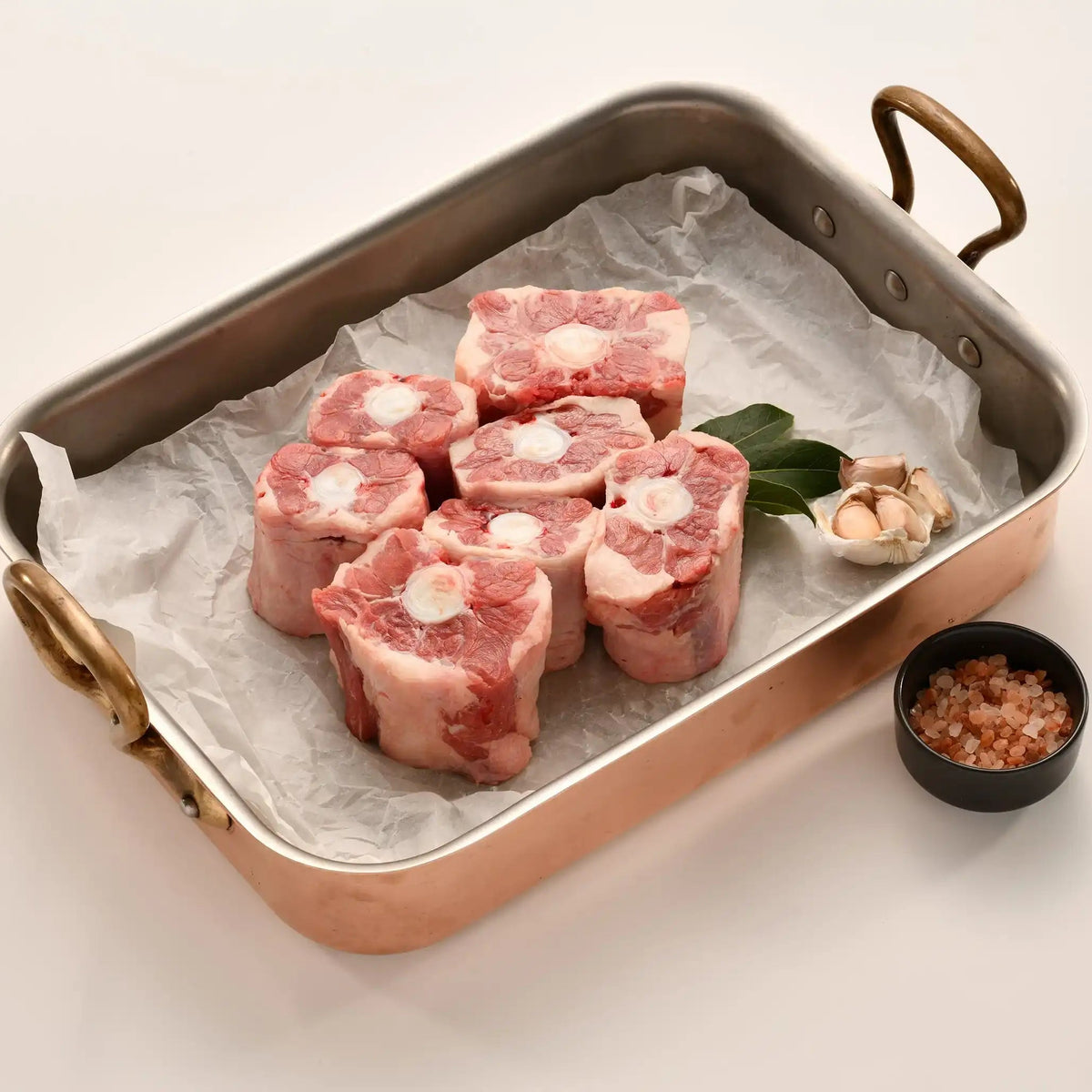 Raw oxtail pieces in a roasting pan with bay leaves, garlic, and pink salt on white background.