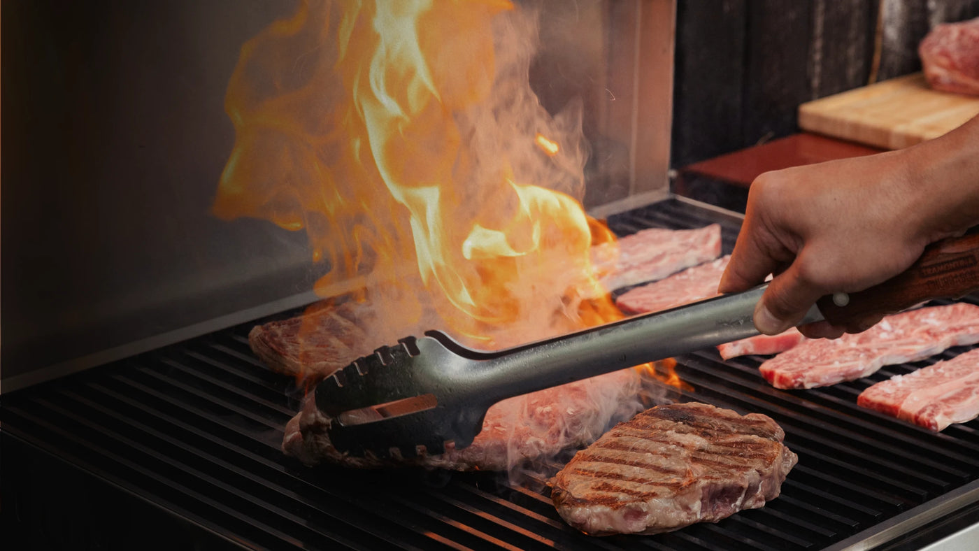 A close-up action shot of a hand using metal tongs to flip a steak with bright orange flames licking the side of the grill and other raw meat