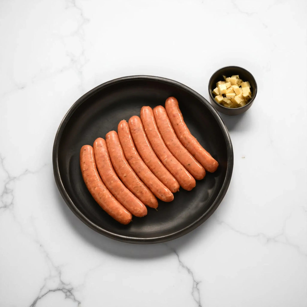 Raw lamb and haloumi sausages arranged on a black plate with a cup of cheese cubes on the side.