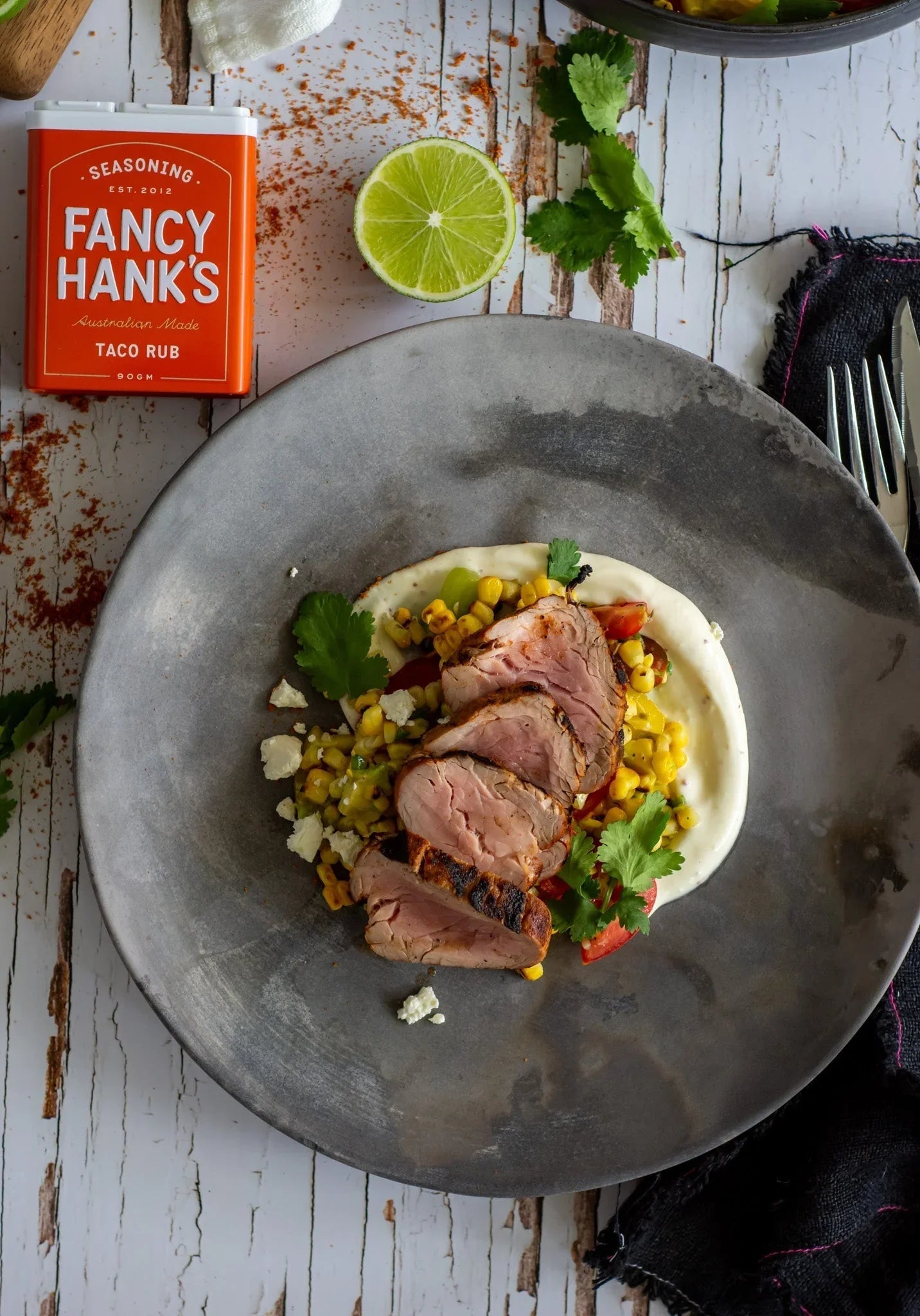 Sliced grilled beef with corn salsa and creamy sauce, garnished with cilantro and lime, featuring Fancy Hank's taco rub