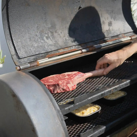 Hand placing premium tomahawk steak on smoker grill with mac and cheese, Peter Augustus quality meats