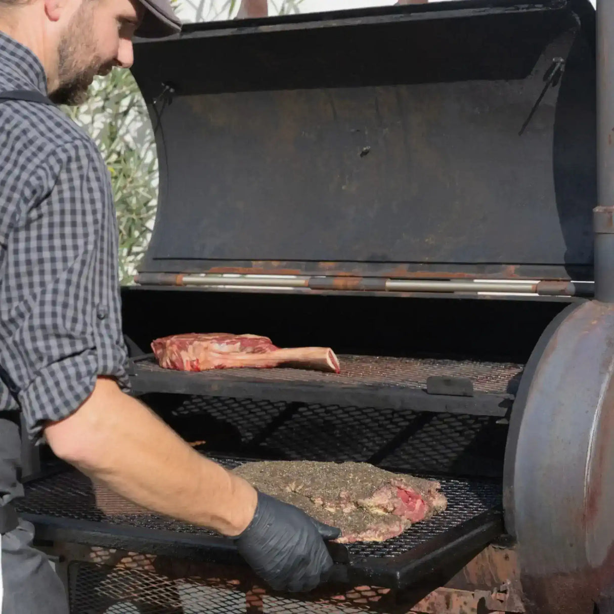 Butcher placing seasoned premium beef cuts, including tomahawk steak, in a smoker grill outdoors