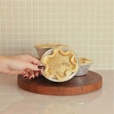 Hand holding a savory meat pie in a foil tin, displayed on a wooden board with more pies