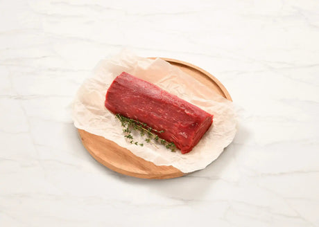 Raw Angus eye fillet log on parchment with fresh herbs on a wooden board, marble background