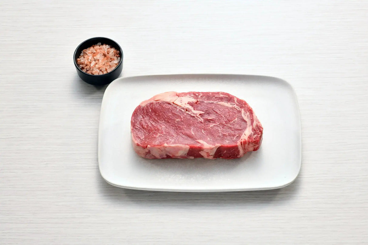 Raw Angus rib fillet steak on white plate with bowl of pink Himalayan salt
