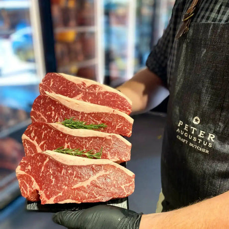 Four Angus striploin steaks with marbling on a tray, held by Peter Augustus Craft Butcher