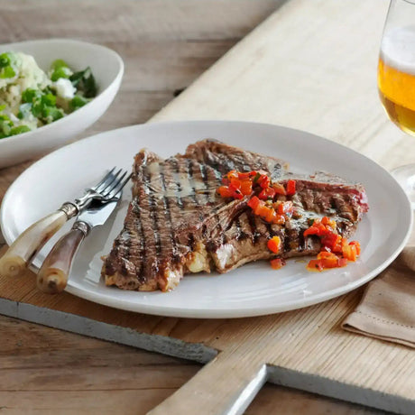 Grilled T-bone steak with diced red peppers, served with salad and beer on a rustic table