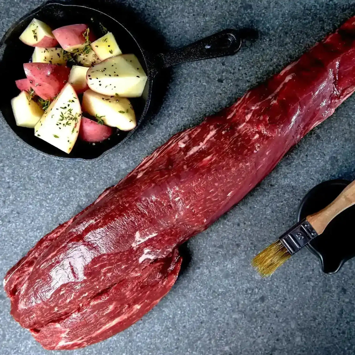 Raw Black Onyx beef tenderloin fillet with diced potatoes and a basting brush on a stone surface