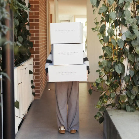 Person carrying three Peter Augustus Craft Butcher delivery boxes in a bright hallway with plants
