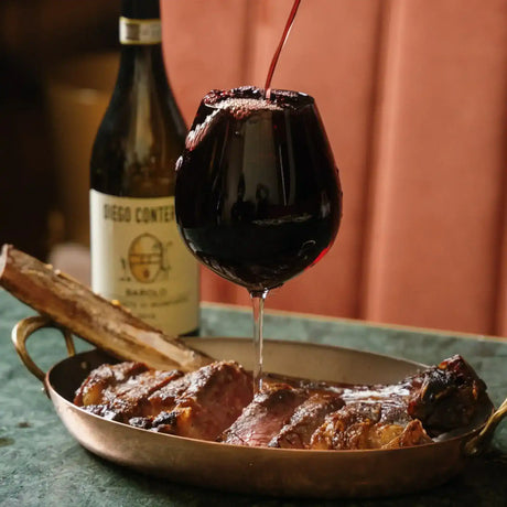 Sliced grilled steak in copper pan with red wine being poured, bottle in background