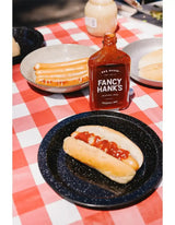 Hot dog with BBQ sauce, sausage rolls, and Fancy Hank's BBQ sauce bottle on picnic table