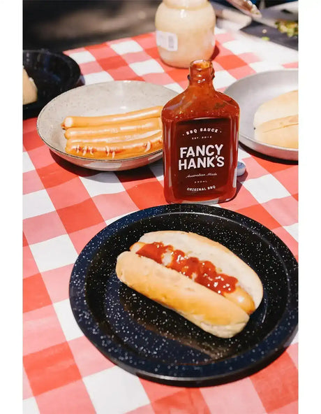Hot dog with BBQ sauce, sausage rolls, and Fancy Hank's BBQ sauce bottle on picnic table