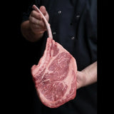 Hand holding a marbled tomahawk steak with visible bone against a dark background