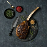 Grilled Wagyu tomahawk steak on a stone board with herbs, spices, and a chef knife