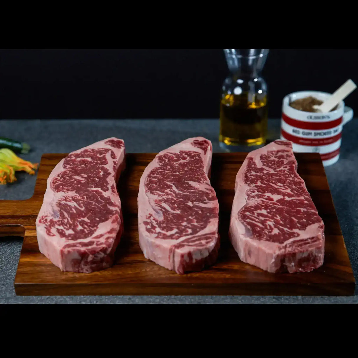 Three marbled beef steaks on wooden board, ready for cooking, with oil and seasoning in background