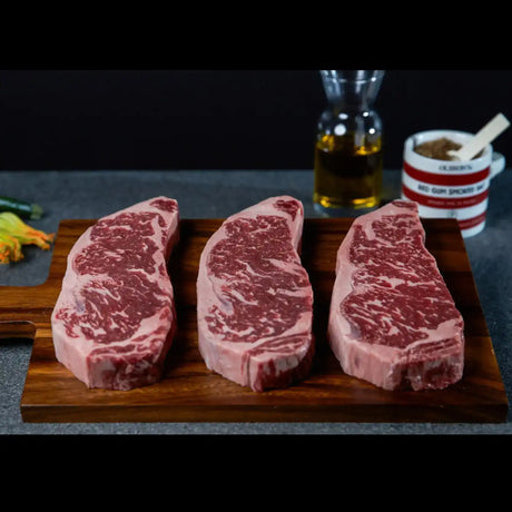 Three marbled beef steaks on wooden board, ready for cooking, with oil and seasoning in background