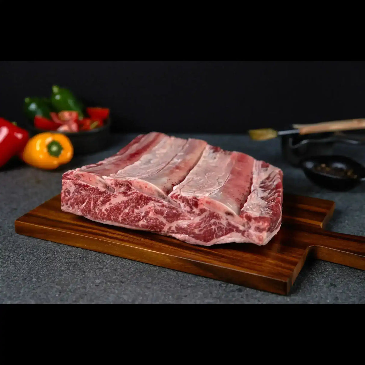 Raw Angus beef short ribs with marbled texture on a wooden board, peppers in background