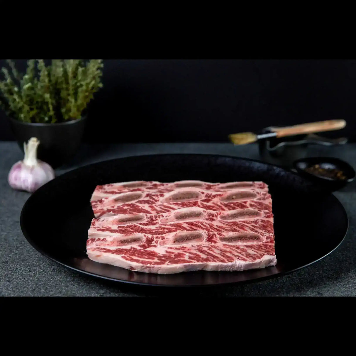 Raw Angus Asado beef short ribs sliced on a black plate, marbled, with garlic and herbs