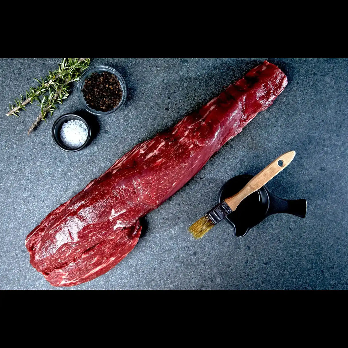 Whole raw Wagyu eye fillet on stone with rosemary, salt, pepper and basting brush