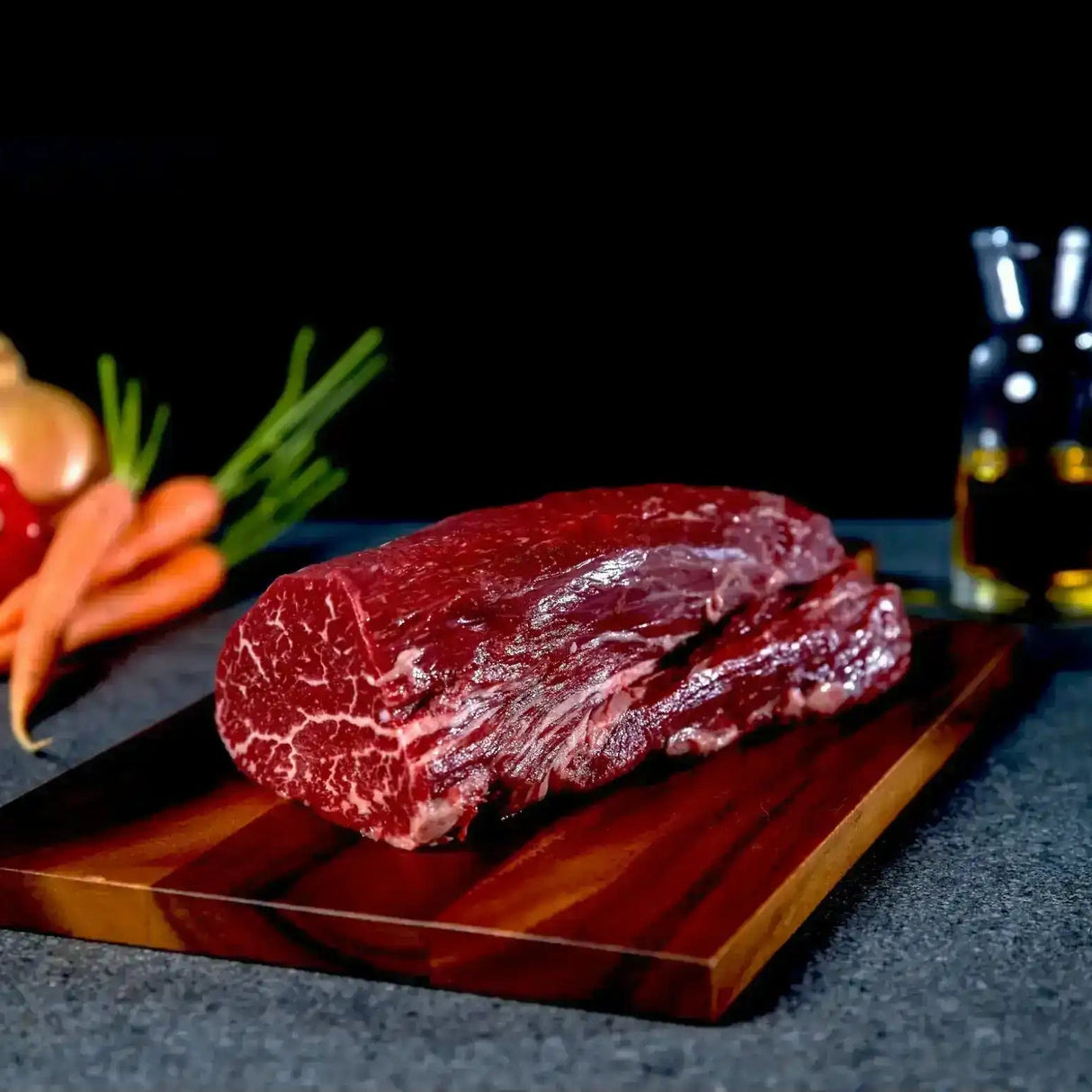 Raw Wagyu eye fillet steak on wooden board with carrots, onion, and oil in the background