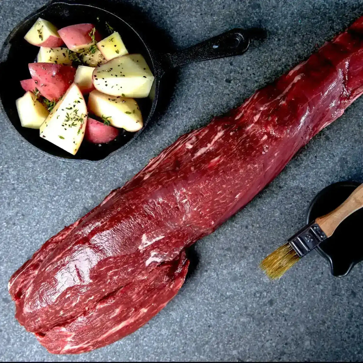 Raw beef tenderloin with potatoes in a skillet and basting brush on a dark stone surface
