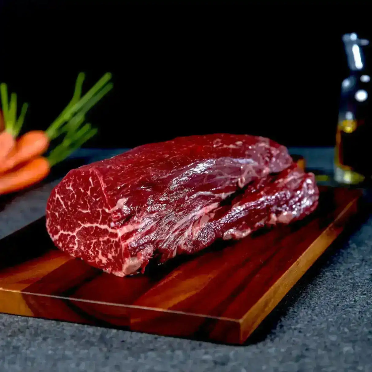Raw marbled beef steak on wooden board with carrots and oil bottle in the background