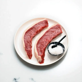 Two raw Angus picanha steaks on a white plate with a bowl of coarse salt