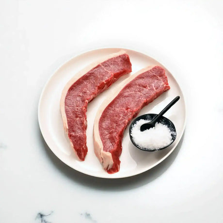Two raw Angus picanha steaks on a white plate with sea salt on a marble surface