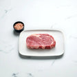 Raw Angus rib fillet steak on a white plate with a bowl of pink salt on a marble surface