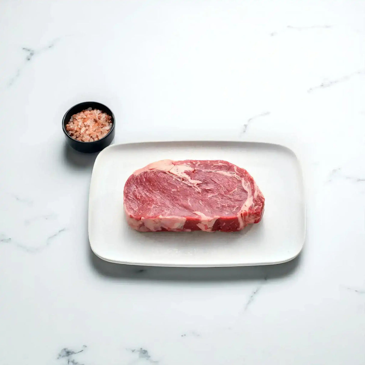 Raw Angus rib fillet steak on a white plate with a small bowl of pink salt on marble background