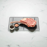 Raw Angus tomahawk steak on wire rack with bowls of black pepper and pink salt on marble background
