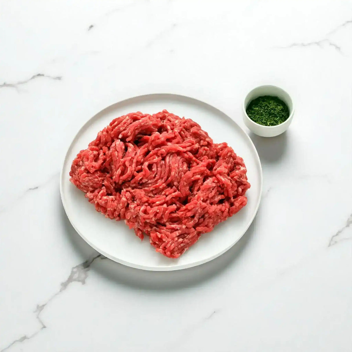 Grass-fed beef mince on a white plate with a small bowl of fresh herbs on a marble surface