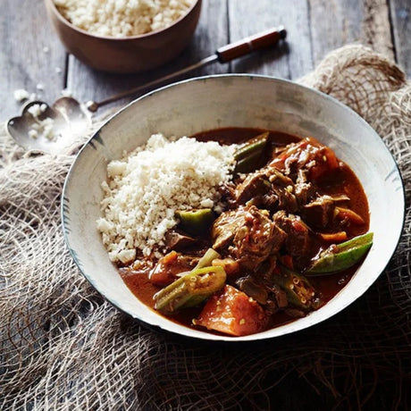 A bowl of chicken and vegetables in a rich brown sauce served with white rice, set on a rustic wooden table with burlap and a small bowl of extra rice in the background.