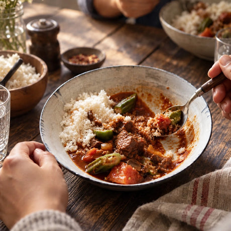 A close-up of a rustic ceramic bowl filled with curry goat meat, green peppers, tomatoes, and white rice, being eaten with a spoon on a dark wooden dining table.