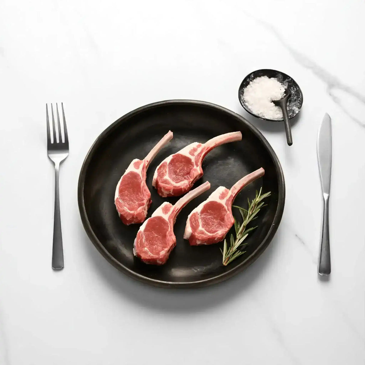 Four raw frenched lamb cutlets on a black plate with rosemary, salt, fork and knife on marble table