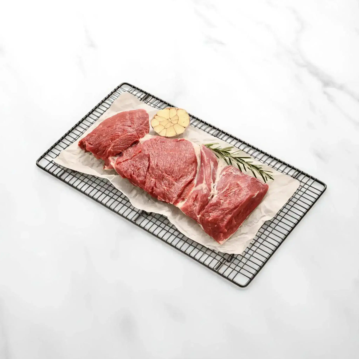 Raw lamb leg butterfly cut on parchment, garnished with garlic and rosemary on wire rack