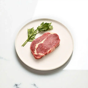 Raw rib fillet steak on a white plate with fresh parsley on marble surface