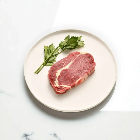 Raw rib fillet steak on a white plate with fresh parsley on marble surface