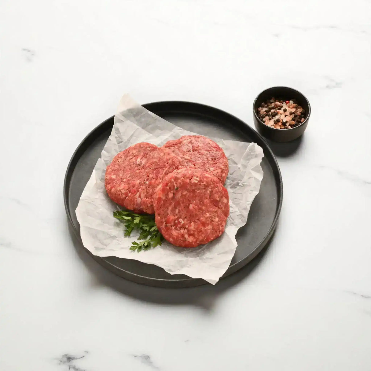 Raw Wagyu beef burger patties on parchment paper with herbs and a bowl of mixed peppercorns
