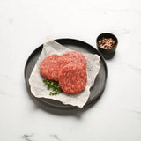 Raw Wagyu beef burger patties on parchment paper with herbs and a bowl of mixed peppercorns