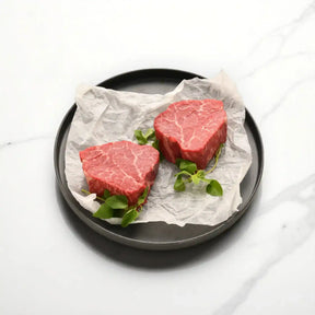 Two raw Wagyu eye fillet steaks with marbling on parchment paper, garnished with greens