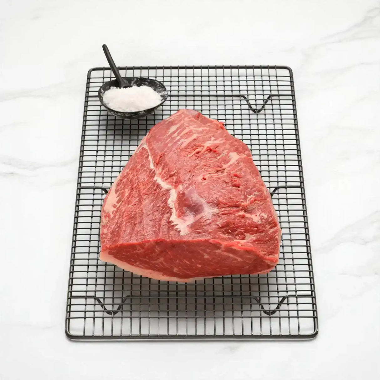 Raw Wagyu rump cap steak on wire rack with a bowl of coarse salt on marble background
