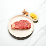 Wagyu sirloin steak with marbling on plate, garlic bulb, and sliced yellow potatoes on marble surface