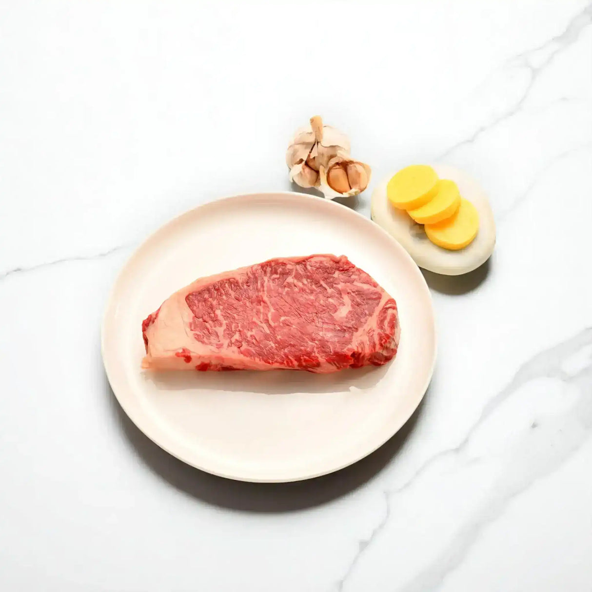 Raw Wagyu sirloin steak on plate with garlic and sliced potatoes on marble background