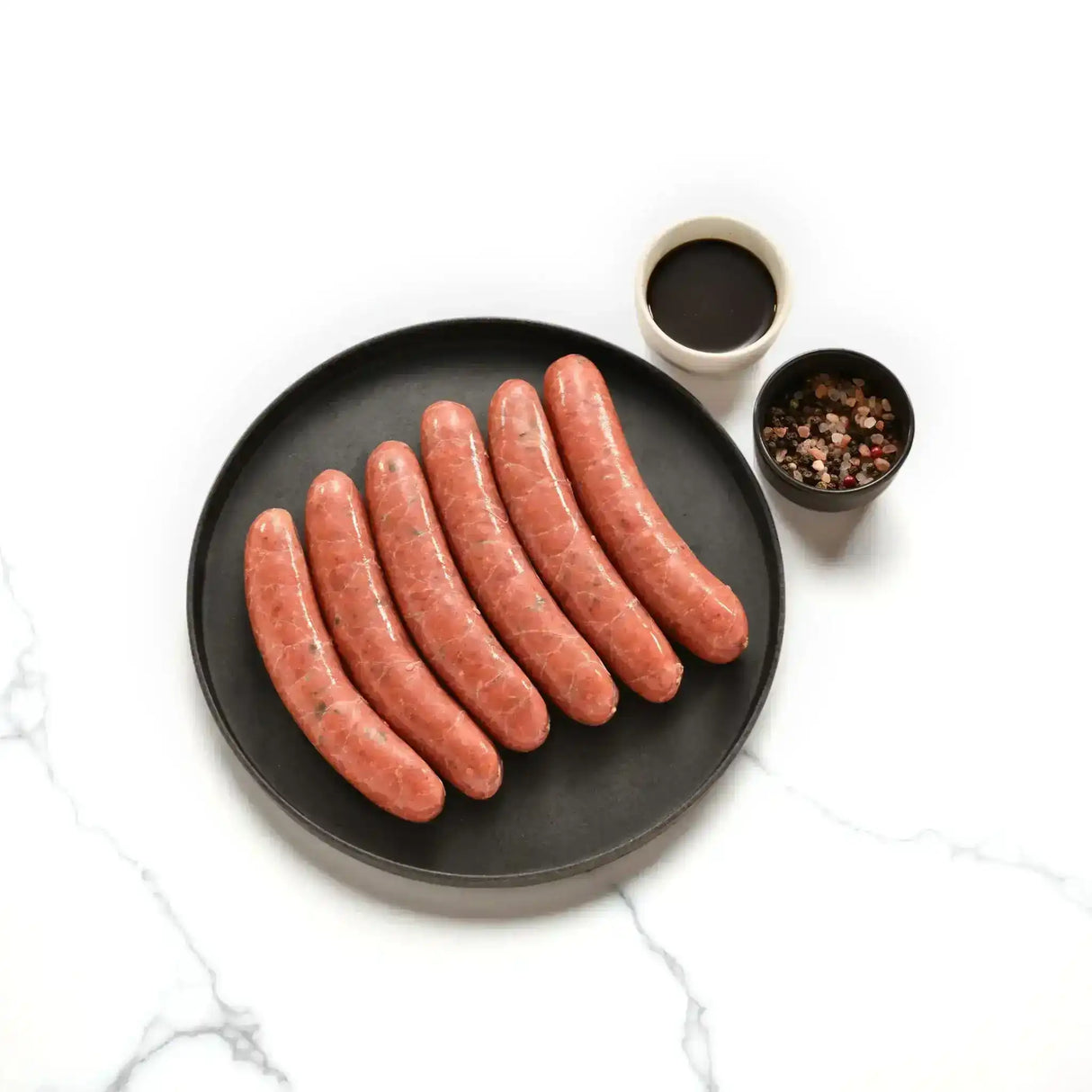 Raw Wagyu beef sausages on black plate with Worcestershire sauce and peppercorns